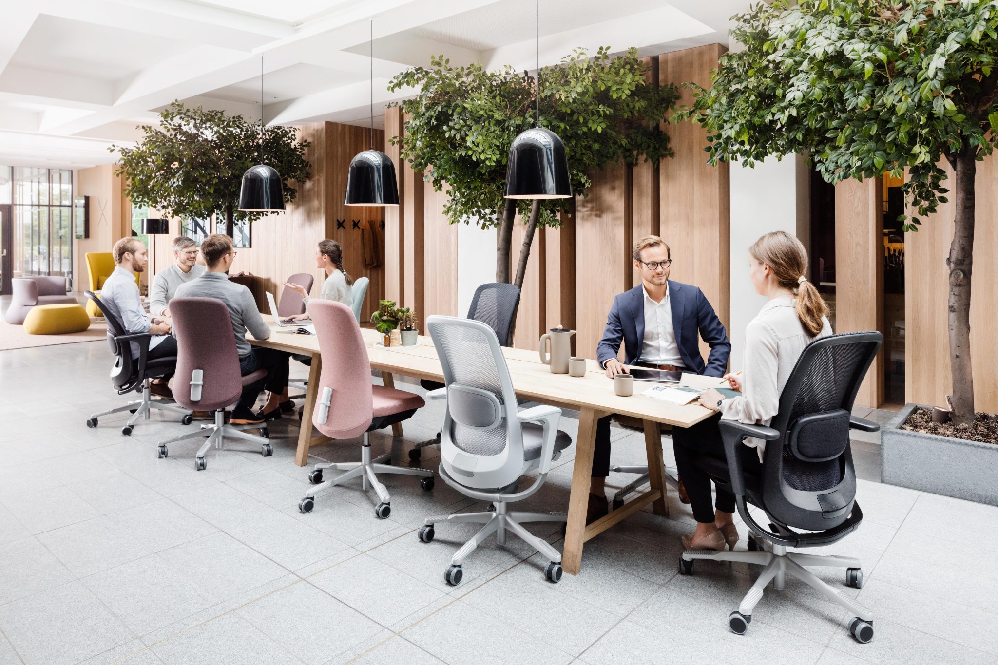 image shows a boardroom table with 8 Hag Sofi chairs or various colours nd styles. 6 people are sat around the table is various levels of discussion and work. the background has wooden slat walls, internal trees, and three large lights above the table, with black lampshades