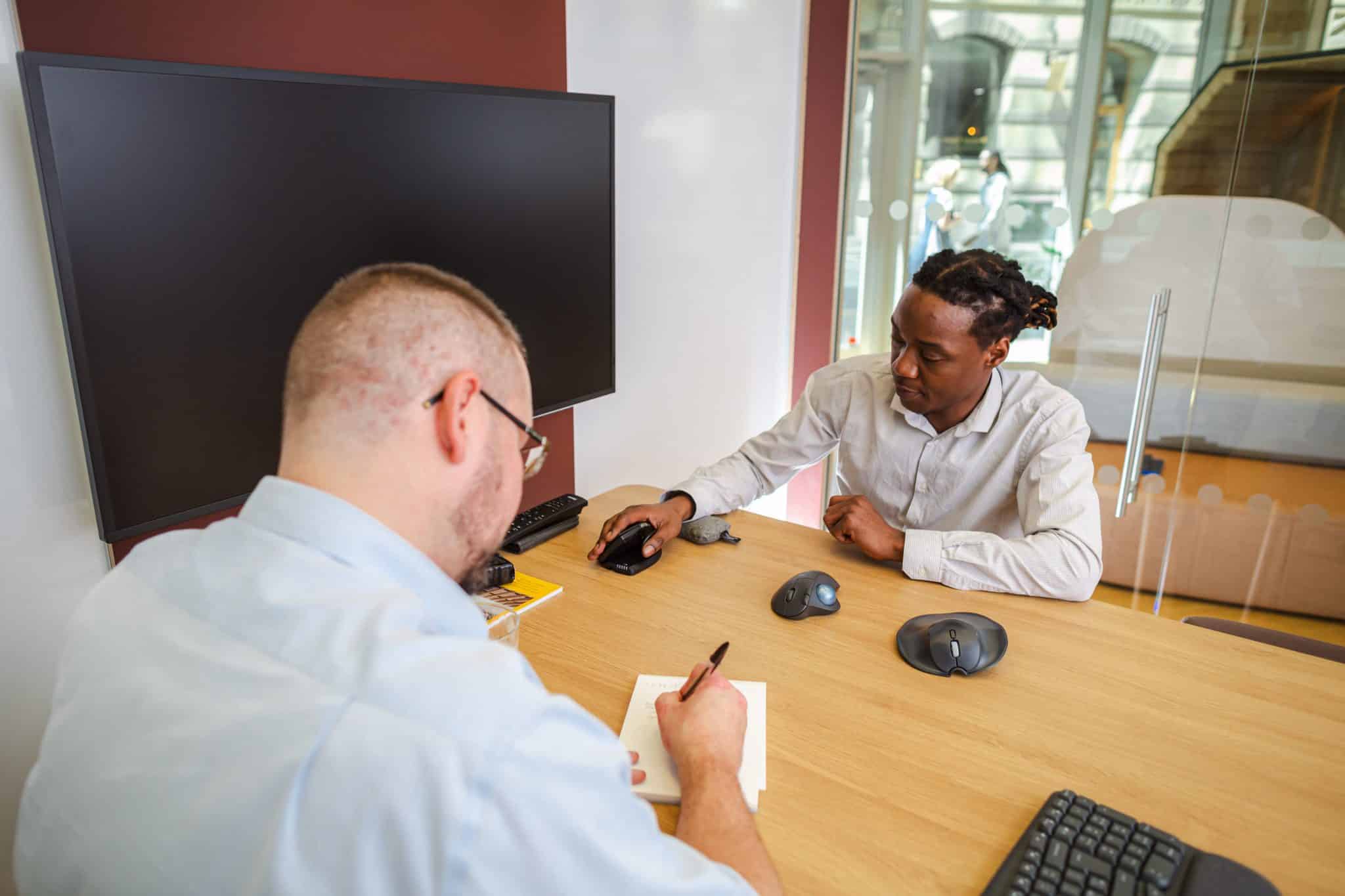 image shows two men in an assessment. several computer mice are being trialled by the man in the background. the man in the foreground is writing notes onto a notepad