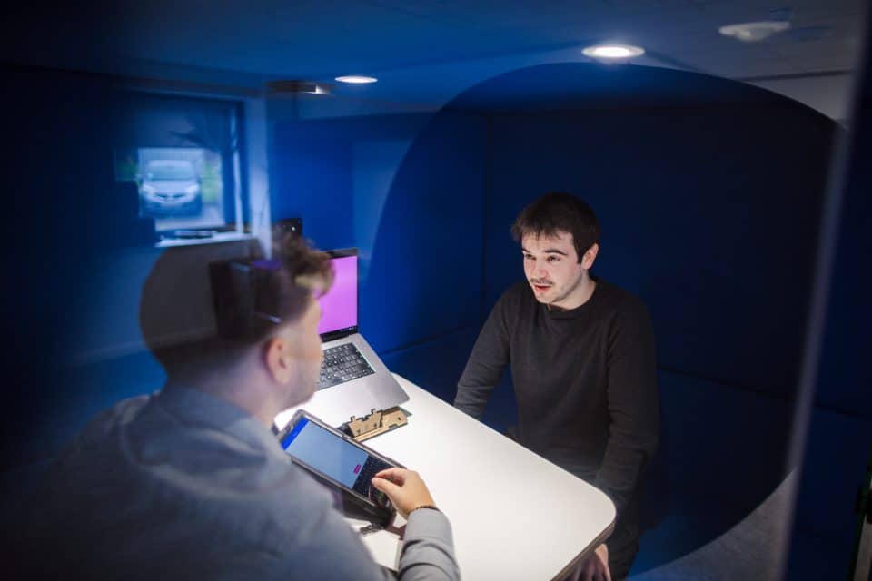 Assistive technology training in Salford showing a face to face coaching discussion where a participant sits inside a blue acoustic pod while a trainer demonstrates assistive technology on a tablet and laptop.