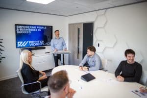 Management presentation taking place in a modern meeting room, with a presenter standing at a lectern and colleagues seated around a table, engaging in discussion and collaboration during a team briefing.