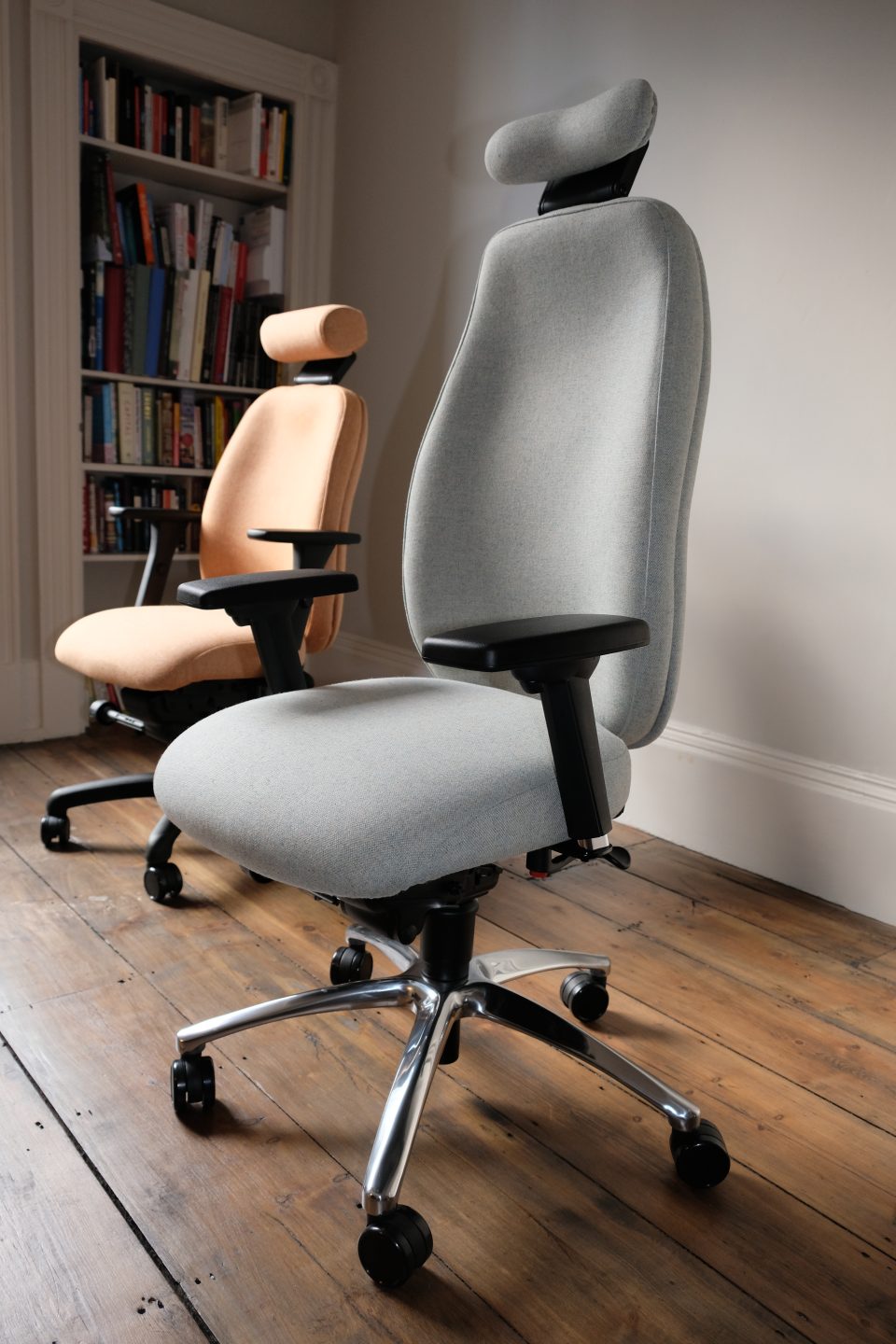 Two Ergochair Adapt 600 ergonomic chair's, one upholstered in light grey, one upholstered in light orange. image shows chair in a home setting, infront of a white bookcase