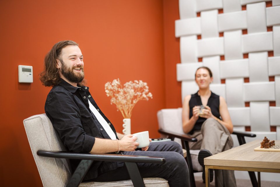 image shows a man and a woman sat on white chairs smiling, in conversation withsomeone off camera. the woman is in the background, holding a tea cup. the walls are orange and cream. a coffee table is in the foreground