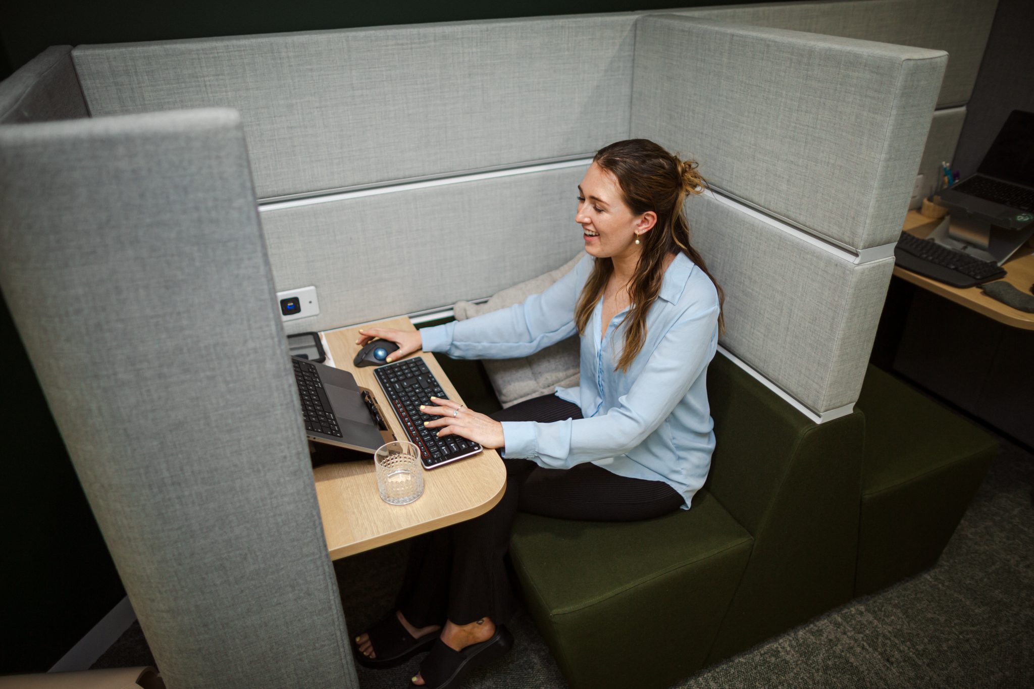 image shows a woman with brown hair and light blue blouse sat in a grey desk pod. she is using a keyboard and mouse separate to her laptop which is raised on a laptop stand