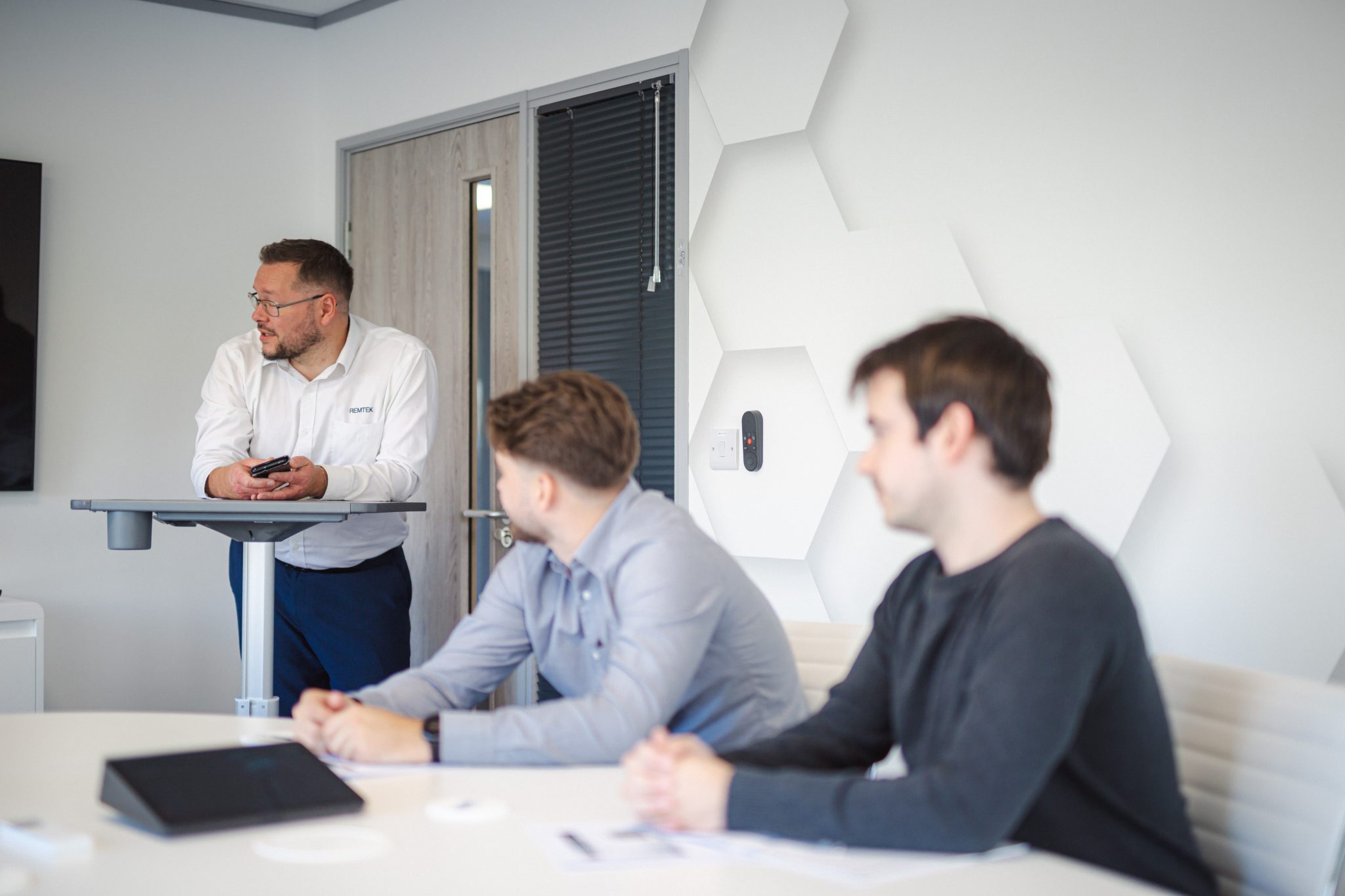 Remtek promotional image showing a presenter standing at a lectern leading a workplace discussion with colleagues seated at a meeting table in a modern office environment.