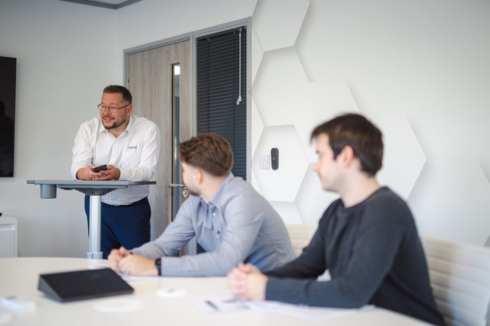 image shows 3 males in a meeting room. one is stood at a podium looking at someone off camera. 2 are sat down looking at the speaker. They seem to be discussing access to work