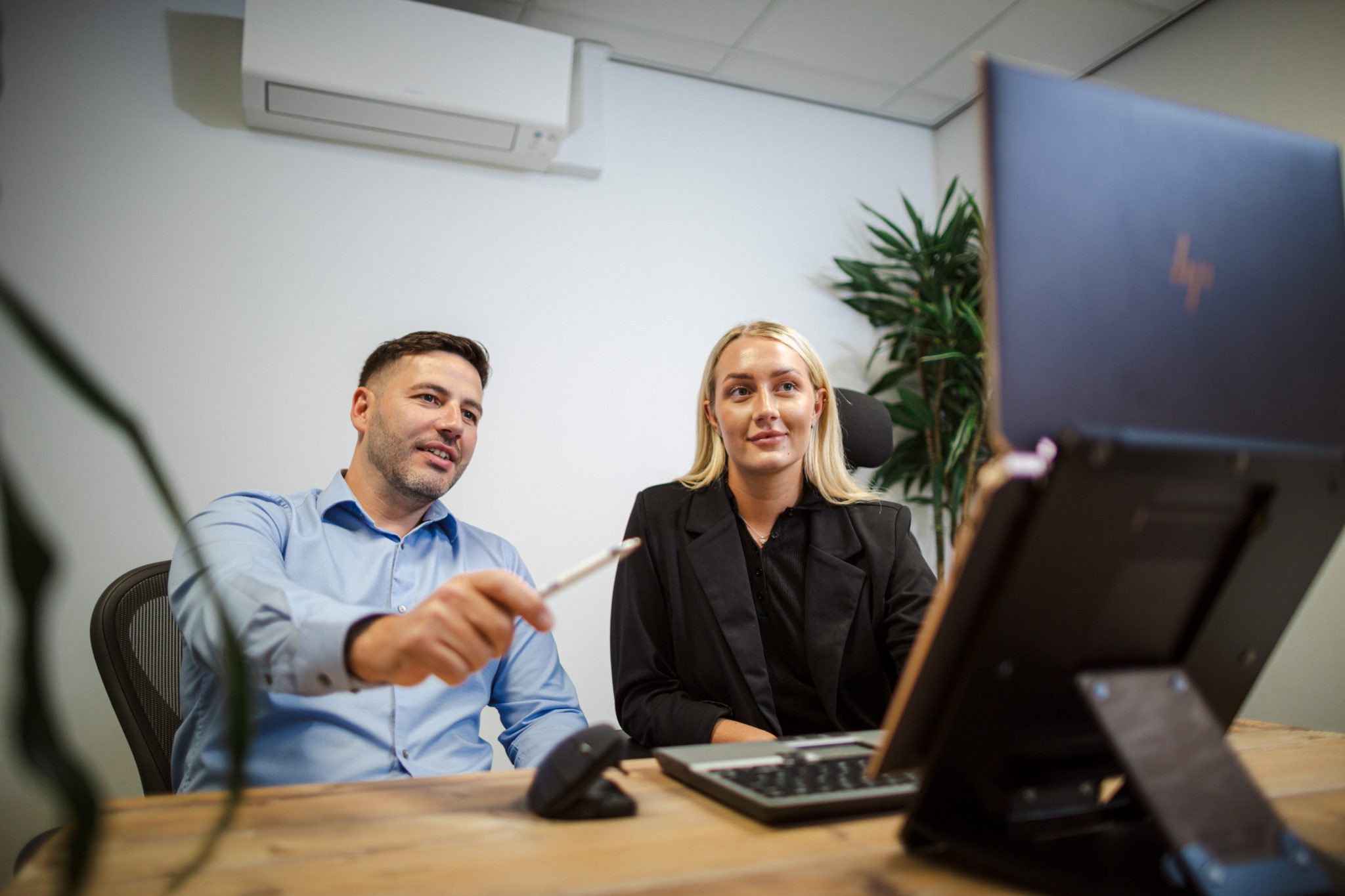 Image shows 2 people sat at a desk looking at a laptop screen. the man on the left wearing a lihght blue shirt, with black hair, he is smiling, and pointing at the screen. The blonde woman in a black business suit is on the right of the image, smiling at what is shown on the screen. there are office plants in the background