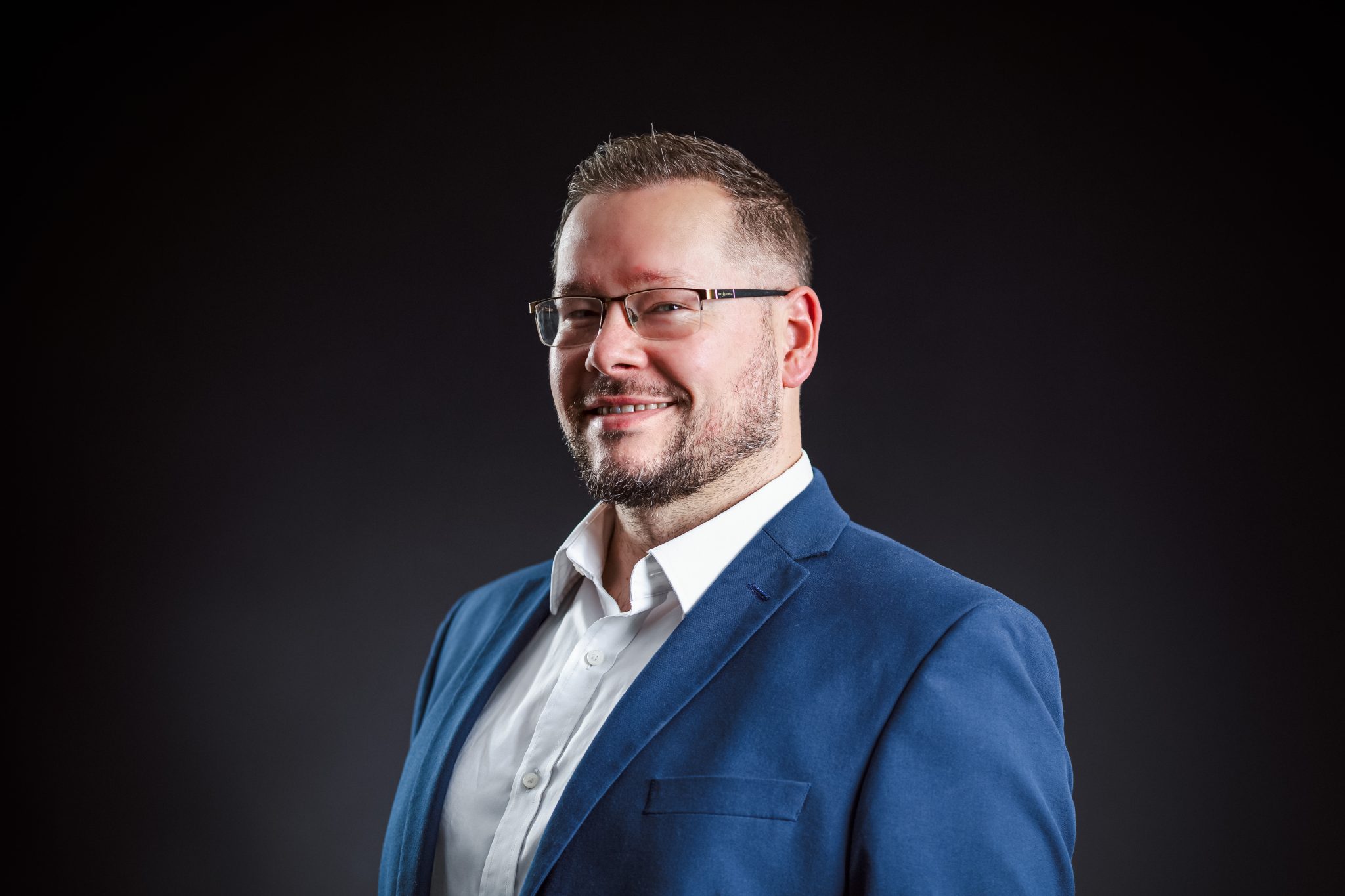 Image is a headshot of Chris Barlow from Remtek Workplace. He has short, brown, styled hair, a neatly trimmed beard and horn rimmed glasses. He is smiling. He is wearing a white shirt and navy blue suit. the image background is black