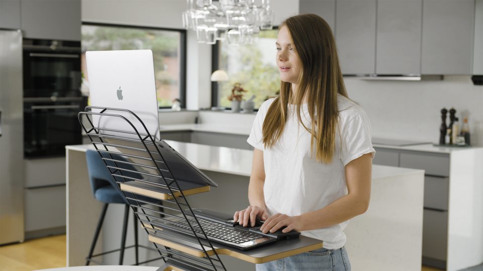 Mousetrapper standfriend in use with a laptop set up. platform is on a wooden desk, with a window showing natural light coming from behind the desk illuminating the workspace. a woman is using the keyboard on the keyboard platform and looking at the laptop screen.