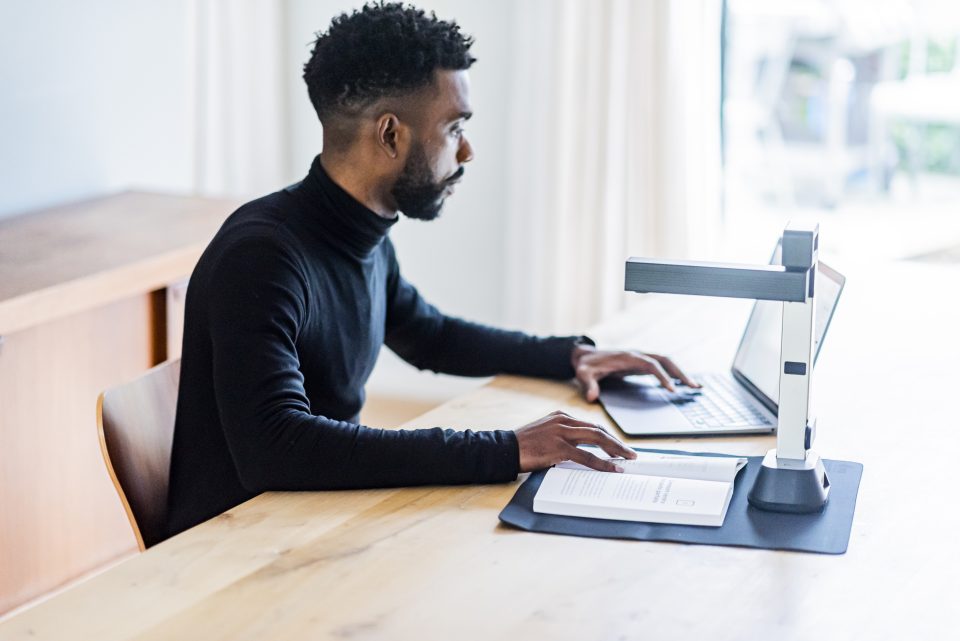 IRIScan Desk 6 set up on a light oak desk, a man in a black turtleneck jumper is scanning a bookscanning