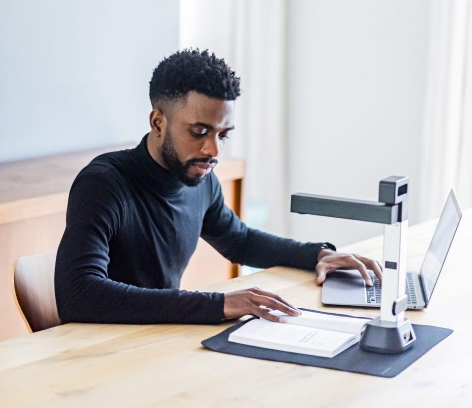 IRIScan Desk 6 set up on a light oak desk, a man in a black turtleneck jumper is scanning a book