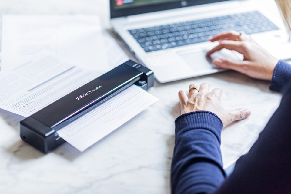 a closeup of the IRIScan Executive 4 Duplex scanning a page of text. a laptop is in the background, and arms in a dark blue jumper are present on the white desk