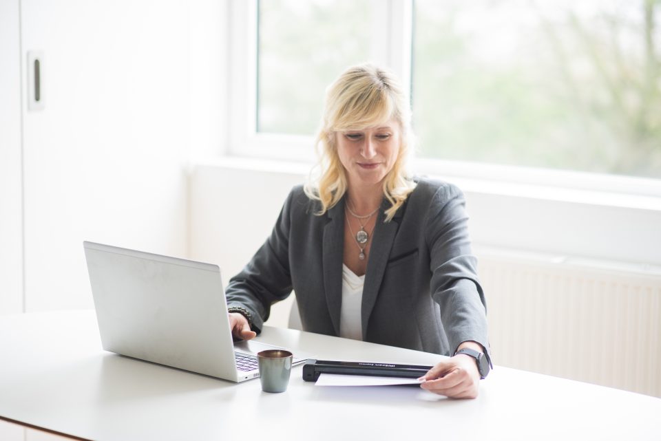 Blonde woman using IRIScan Executive 4 device scanning a page of text she has a laptop and coffe cup on the white desk. a large window with dnatural light is behind her