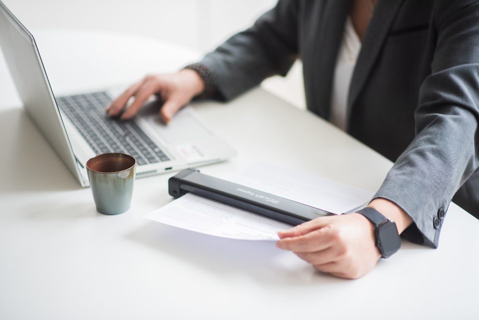 Close up of an office worker using IRIScan Executive 4 device scanning a page of text she has a laptop and coffe cup on the white desk. a large window with natural light is behind her