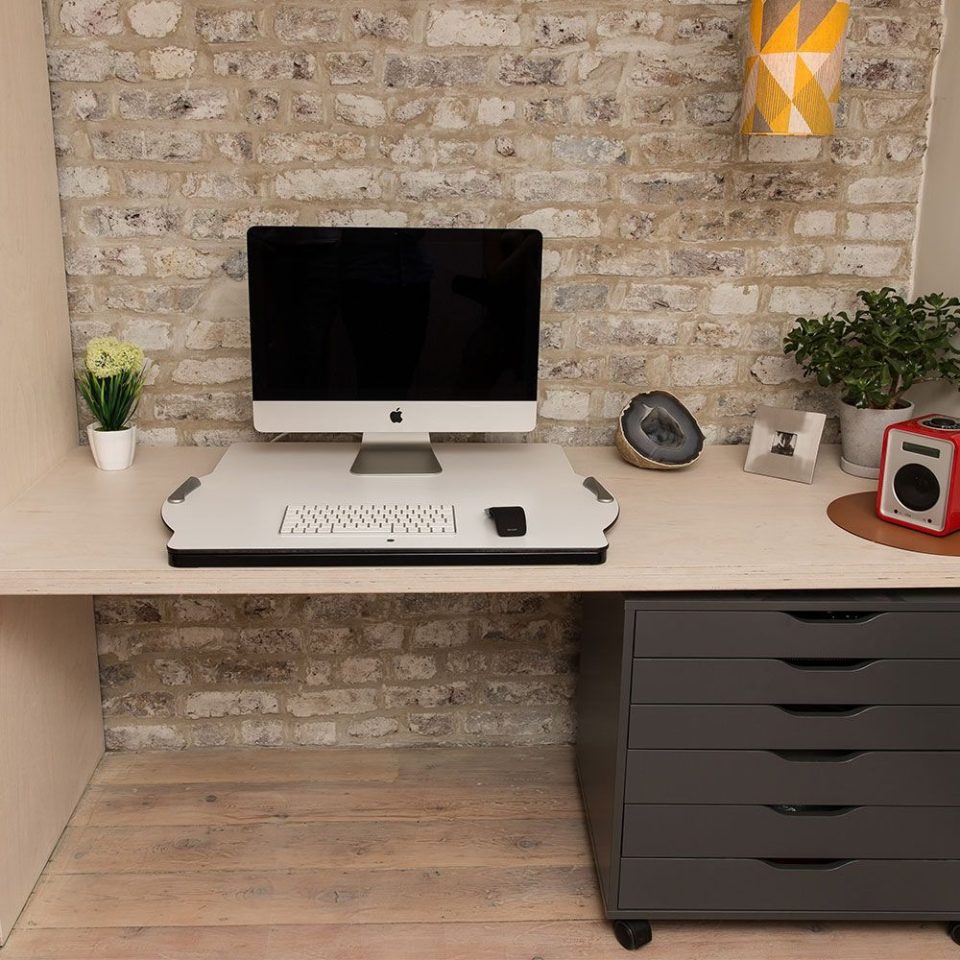 Opløft Sit-Stand Platform in the low position with a monitor, keyboard and mouse present on the platform. platform is on top of a desk which has drawers to the right side of it. the wall behind is grey bricks