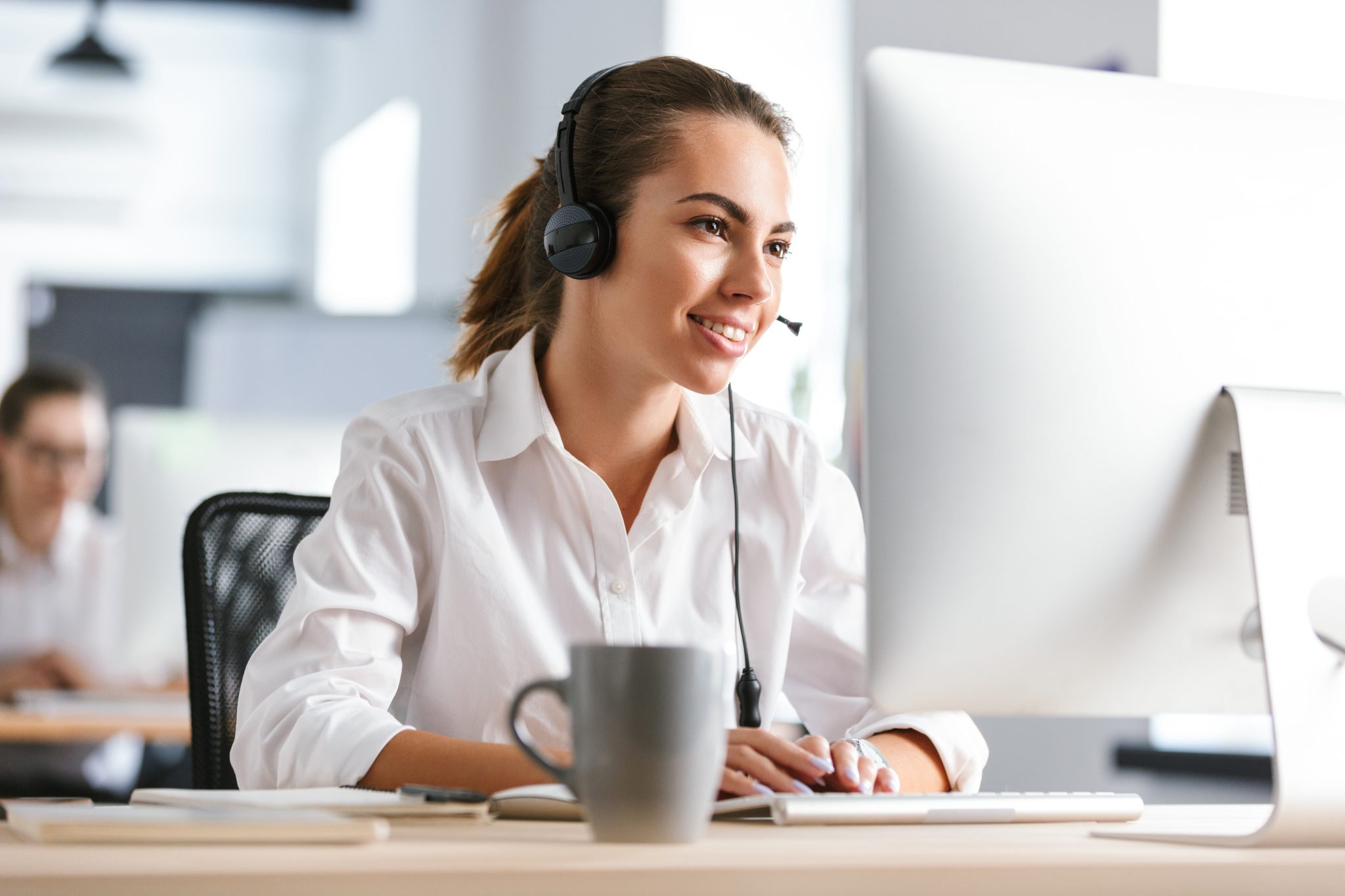 Woman looking at a computer screen. she is wearing a headset, and smiling at the screen. a grey coffee cup is on the desk