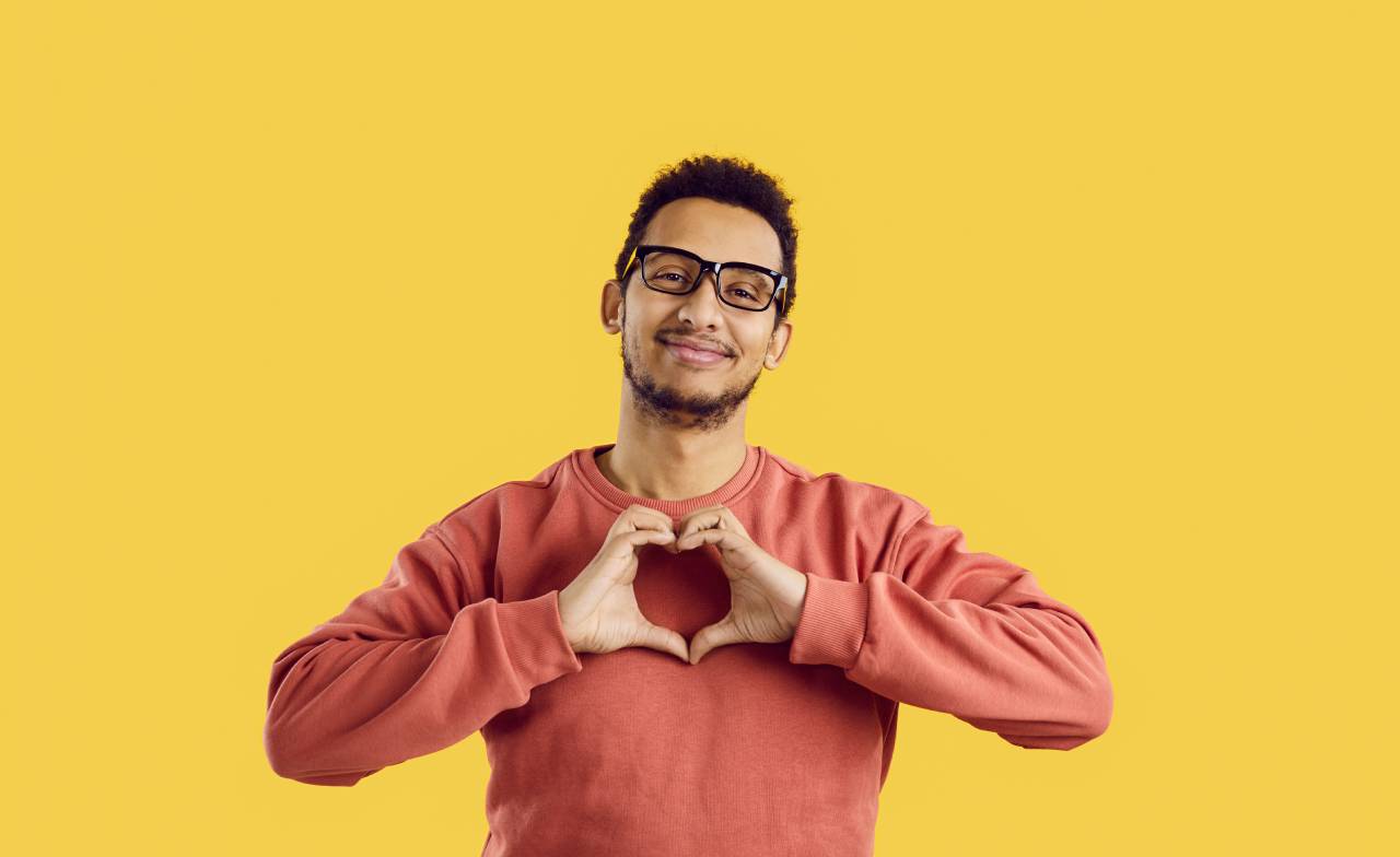 Smiling man wearing glasses forming a heart shape with his hands against a yellow background, expressing positivity, inclusion, and emotional wellbeing through a clear hand gesture.