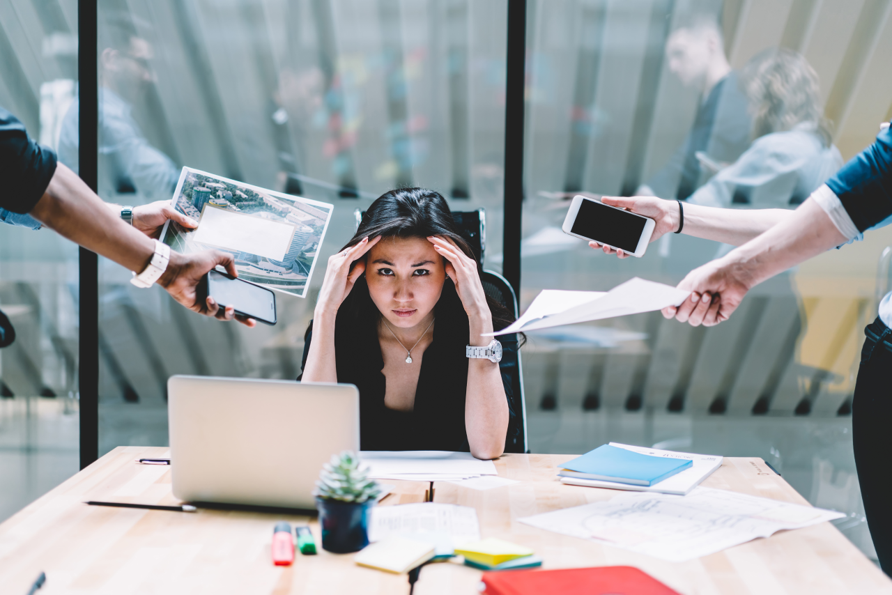 Stressed woman sitting at a desk with her head in her hands, surrounded by colleagues holding out phones, papers and a tablet, symbolising workplace overload and pressure.