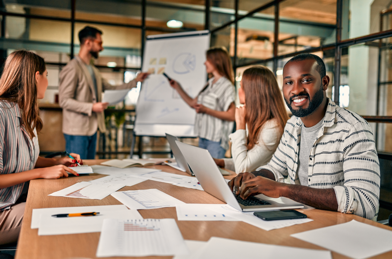 Group of colleagues collaborating in a modern office, with one person smiling at the camera while others discuss charts at a flipchart, representing teamwork and productivity