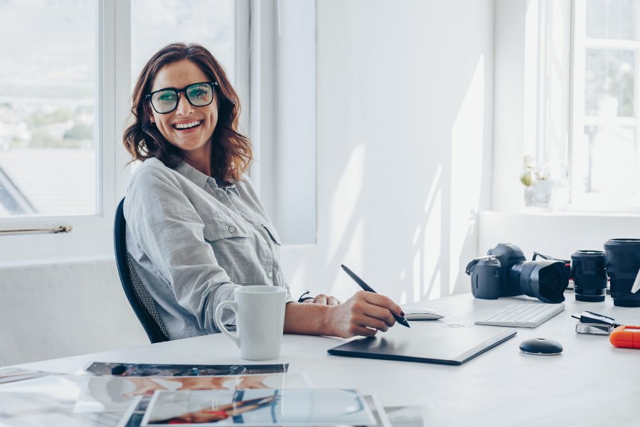 Smiling professional sitting at a bright, organised workstation with computer and camera equipment, representing good display screen equipment use.