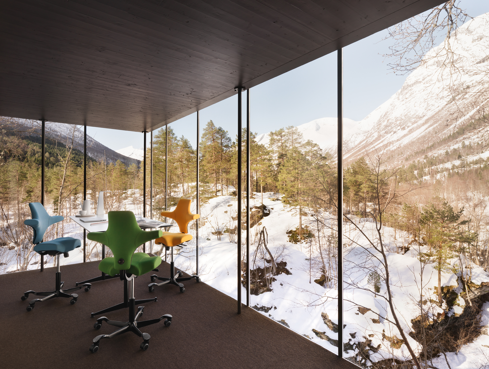A modern workspace with floor-to-ceiling glass walls overlooking a snowy mountain landscape. Inside the room are colorful HÅG Capisco ergonomic chairs in blue, green, yellow, and orange, arranged around a white height adjustable desk.