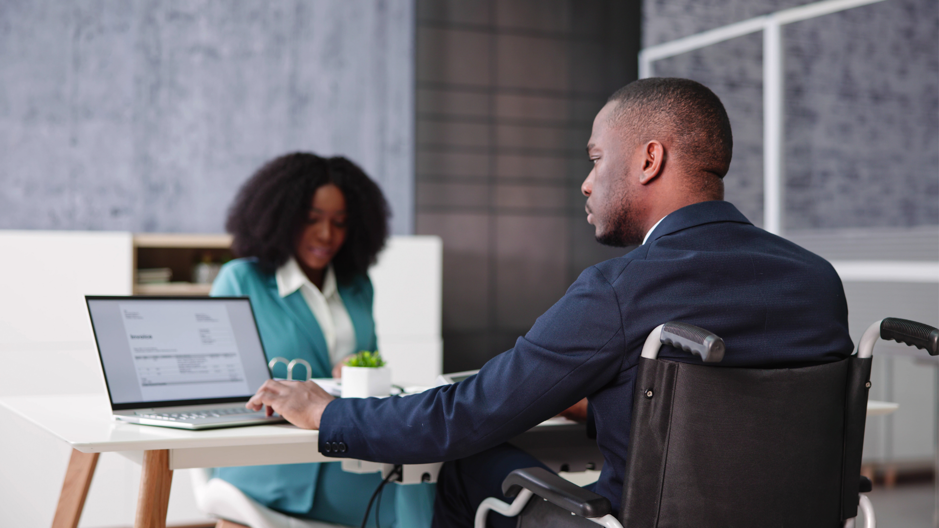 A man in a wheelchair sits at a desk alongside a woman, engaged in conversation or collaboration.
