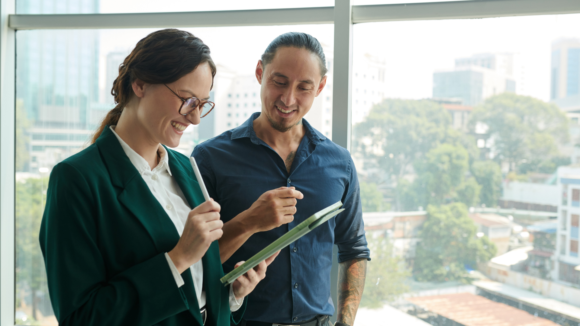 Two business professionals stand by a window, examining a tablet together, engaged in discussion.