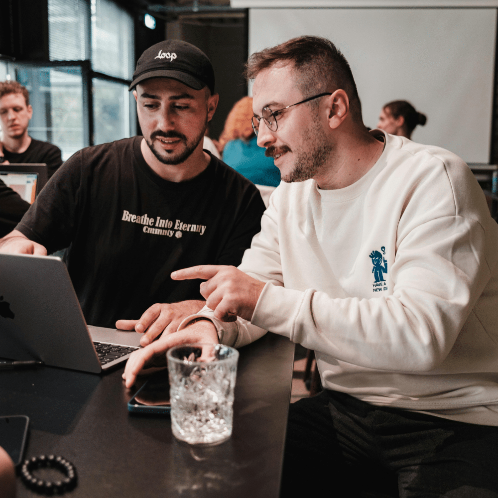 Two men working together on a laptop in a busy collaborative workspace.