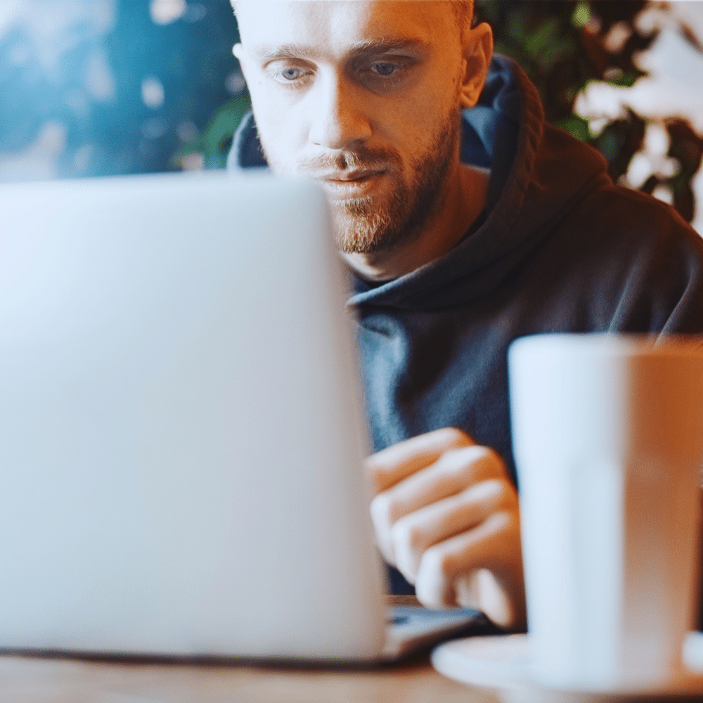 Man concentrating on a laptop at a desk, with a white mug in the foreground.