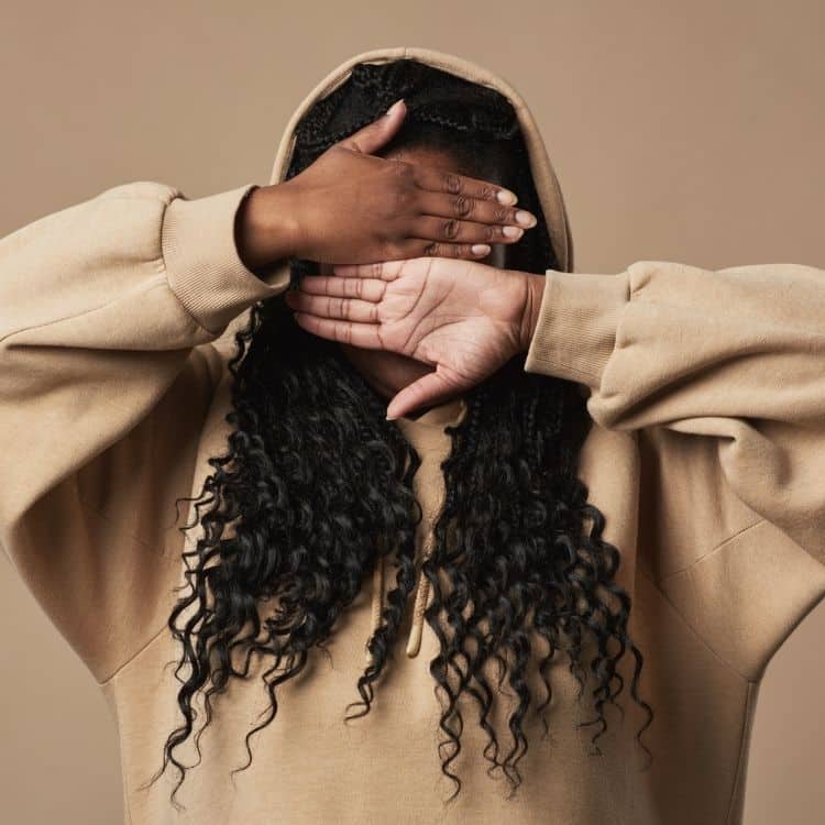 Person hiding oneself by covering their face with both hands while wearing a beige hoodie.