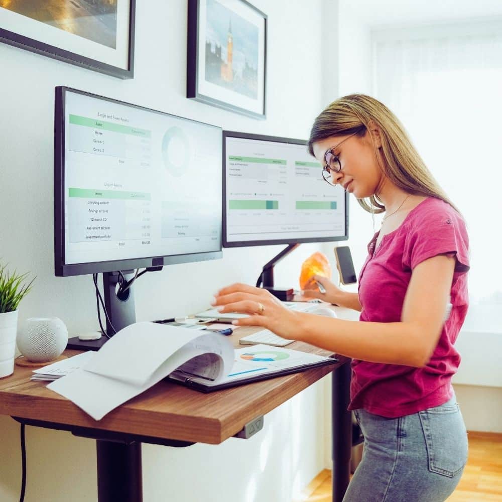 Woman working at a height-adjustable standing desk with dual monitors while reviewing printed documents.