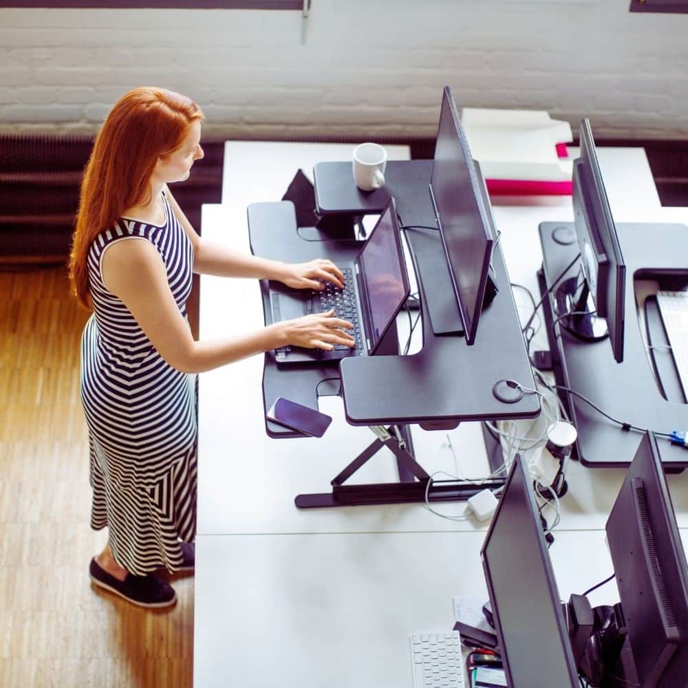 Woman working at a standing desk converter with dual monitors and a laptop in an office.