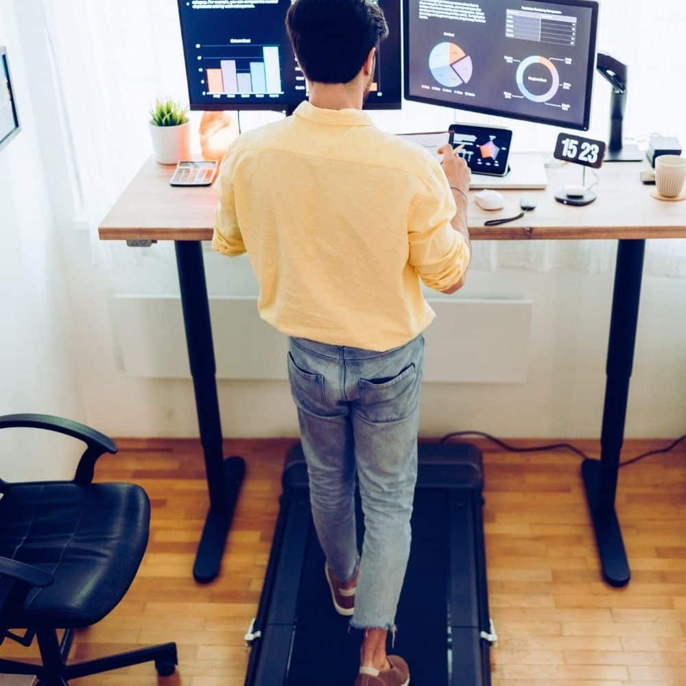 Man walking on an under-desk treadmill while working at a height-adjustable standing desk with dual monitors.