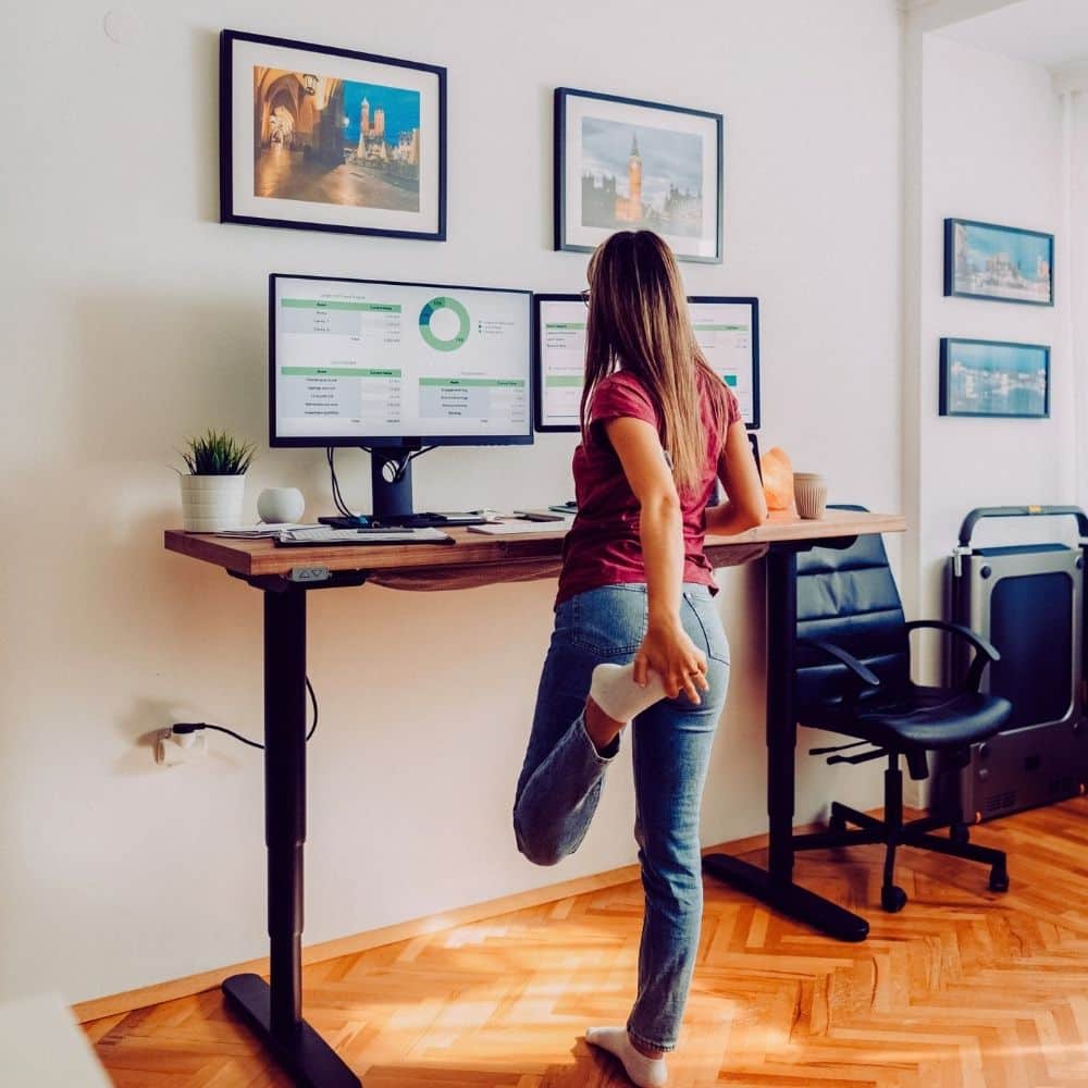 Woman stretching beside a height-adjustable standing desk with dual monitors in a home office.